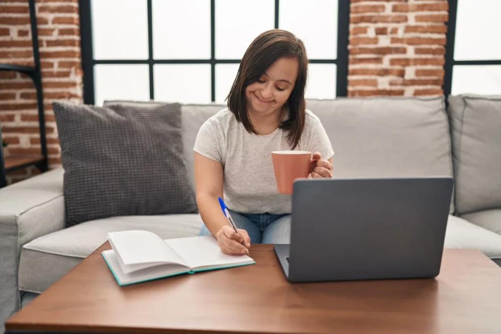 Disabled girl with down syndrome sitting happily at coffee table using laptop and writing in note pad